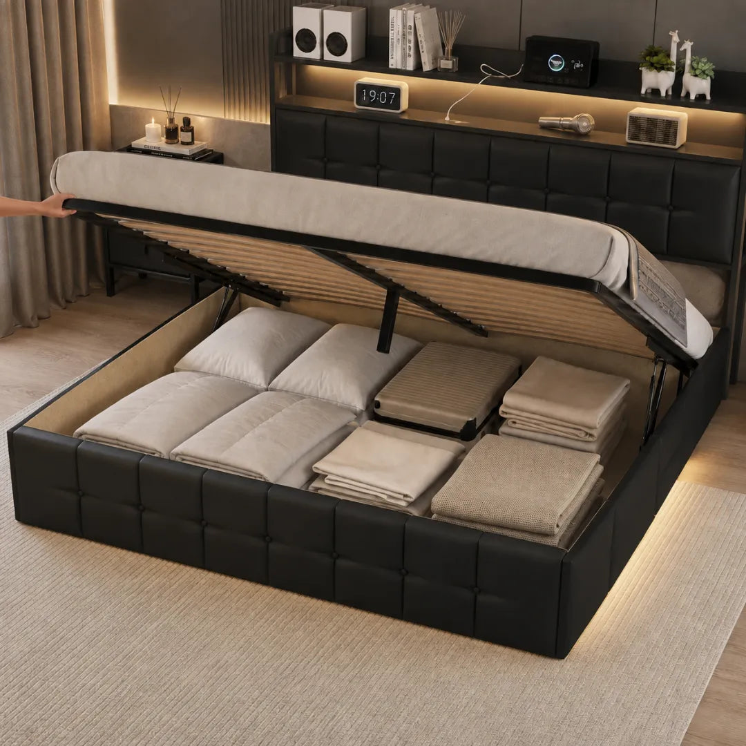 Coffre du lit ouvert d'une main sur son vérin pneumatique, révélant oreillers, linge plié et valise rangés sous le couchage