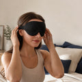 Femme ajustant son masque de nuit Somnolia noir, assise sur un lit aux coussins bleu nuit et lin